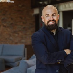 FENBRO SPÓŁKA Z OGRANICZONĄ ODPOWIEDZIALNOŚCIĄ - Portret uśmiechniętego mężczyzny z brodą w granatowym garniturze, z rękami skrzyżowanymi na tle ceglanej ściany i nowoczesnego wnętrza biurowego.