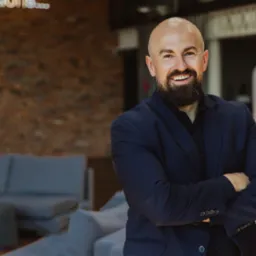 Portret uśmiechniętego mężczyzny z brodą w granatowym garniturze, z rękami skrzyżowanymi na tle ceglanej ściany i nowoczesnego wnętrza biurowego.