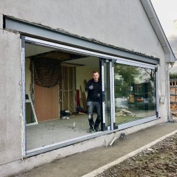 FENBRO SPÓŁKA Z OGRANICZONĄ ODPOWIEDZIALNOŚCIĄ - Montaż dwuskrzydłowych drzwi przesuwnych z szeroką ramą, widoczny pracownik budowlany z podniesionym kciukiem, surowy stan wykończenia wnętrza i elewacji.