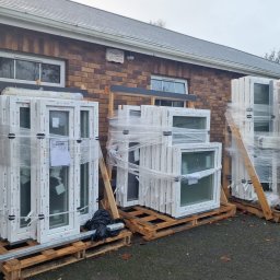 FENBRO SPÓŁKA Z OGRANICZONĄ ODPOWIEDZIALNOŚCIĄ - Pakiety białych okien na drewnianych paletach, zabezpieczone folią stretch, składowane przed budynkiem z cegły.