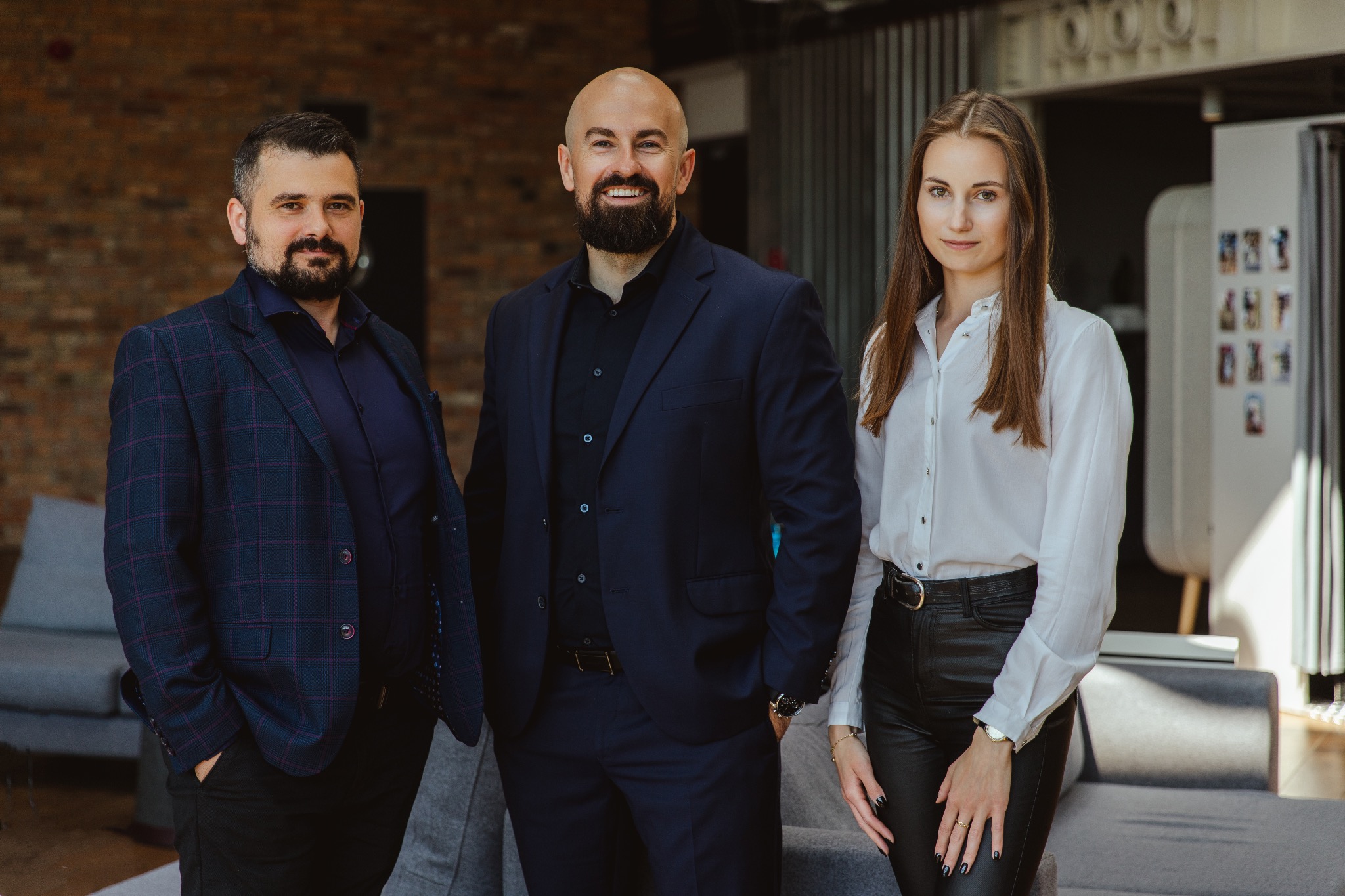 Portret zespołu: mężczyzna w kraciastej marynarce, łysy mężczyzna z brodą w granatowym garniturze i kobieta w białej koszuli i skórzanych spodniach, stojący w nowoczesnym biurze z ceglaną ścianą...