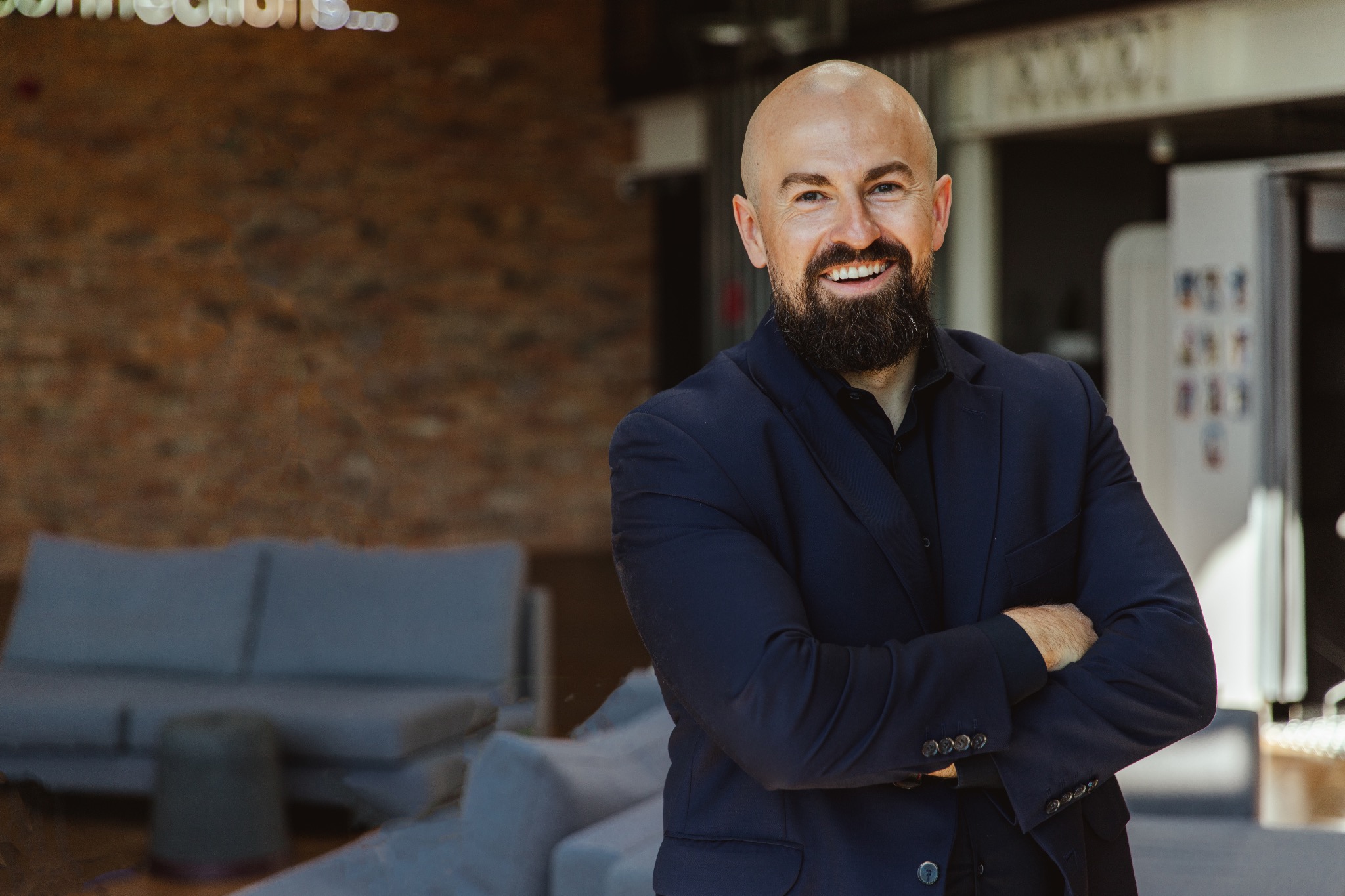 Portret uśmiechniętego mężczyzny z brodą w granatowym garniturze, z rękami skrzyżowanymi na tle ceglanej ściany i nowoczesnego wnętrza biurowego.
