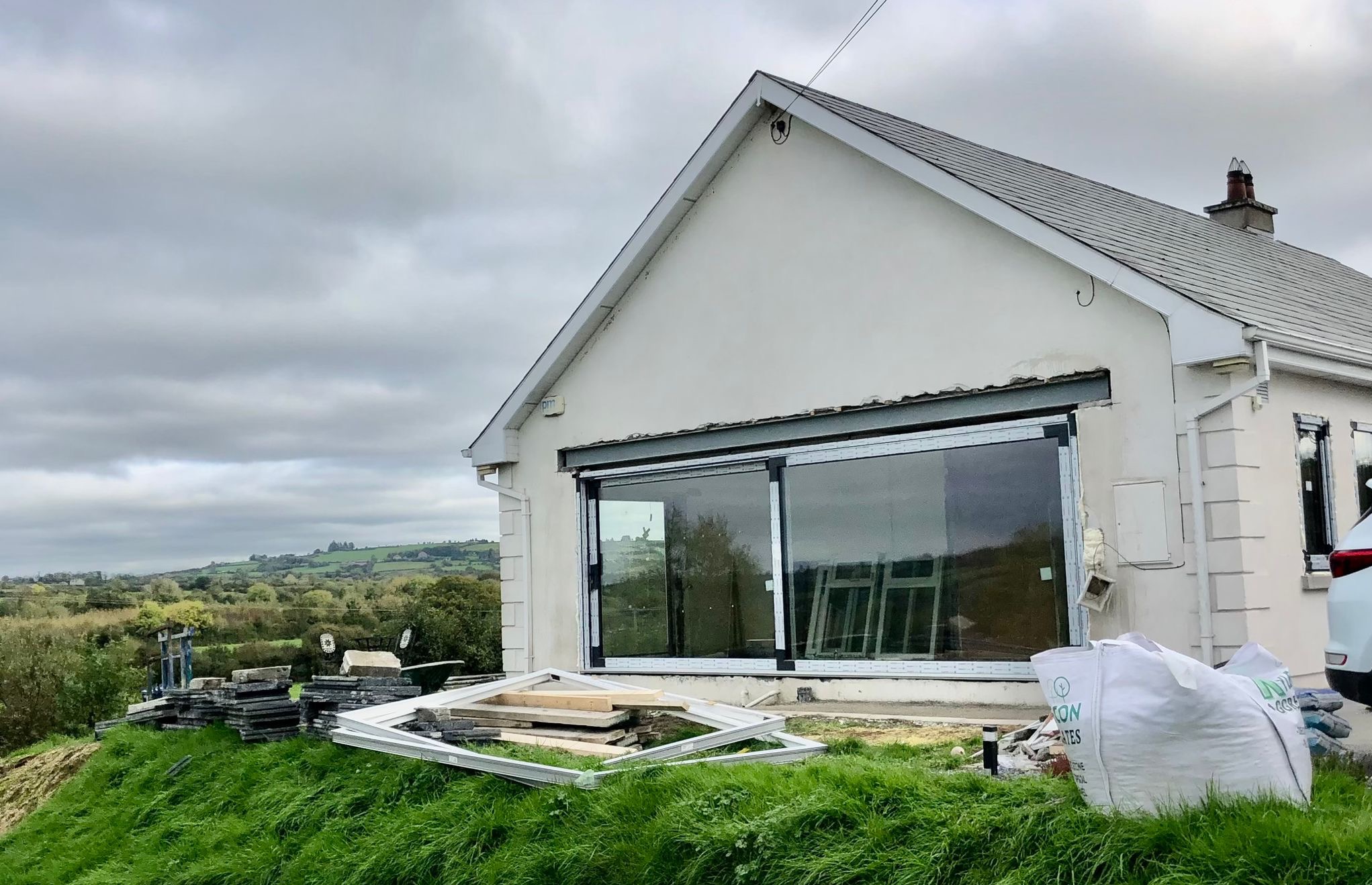 Montaż okna przesuwnego w białym domu jednorodzinnym, widoczne materiały budowlane i elementy okienne na trawie, krajobraz w tle.