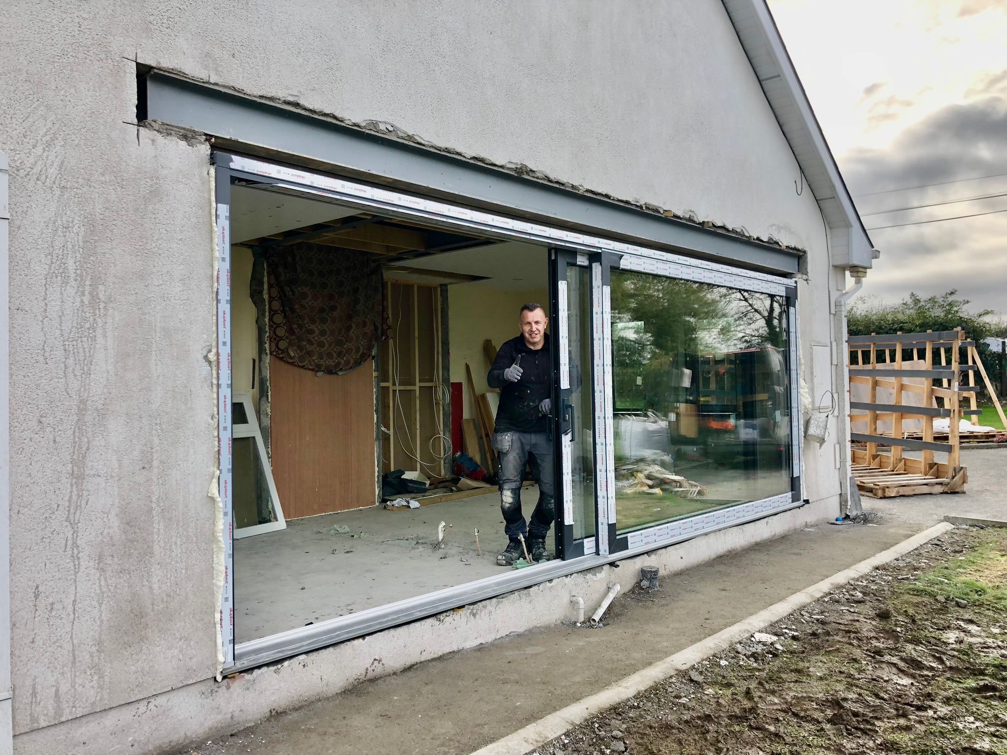 Montaż dwuskrzydłowych drzwi przesuwnych z szeroką ramą, widoczny pracownik budowlany z podniesionym kciukiem, surowy stan wykończenia wnętrza i elewacji.