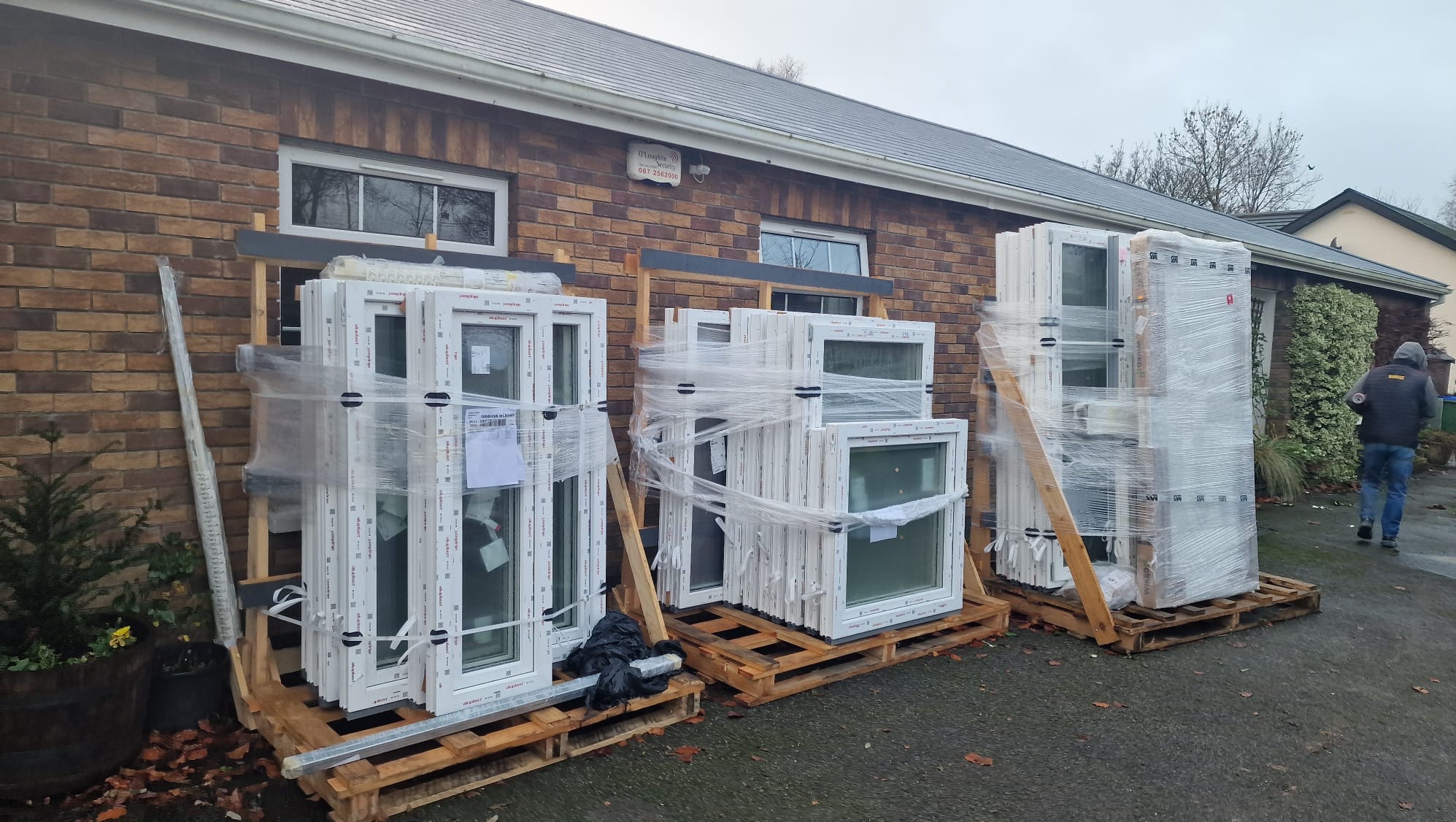 Pakiety białych okien na drewnianych paletach, zabezpieczone folią stretch, składowane przed budynkiem z cegły.