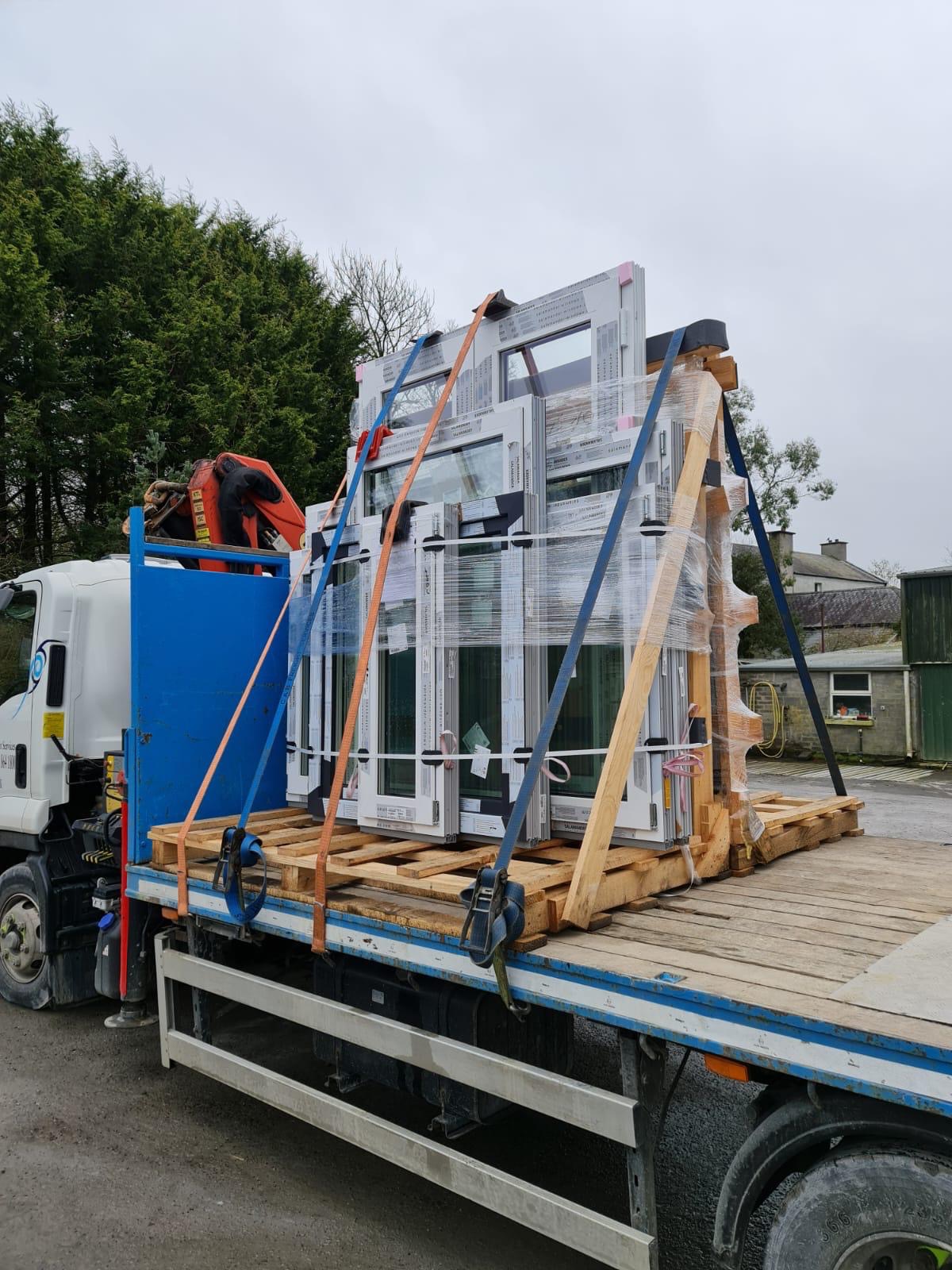Transport okien na paletach, zabezpieczonych folią i pasami, na platformie ciężarówki z dźwigiem, widok z boku.