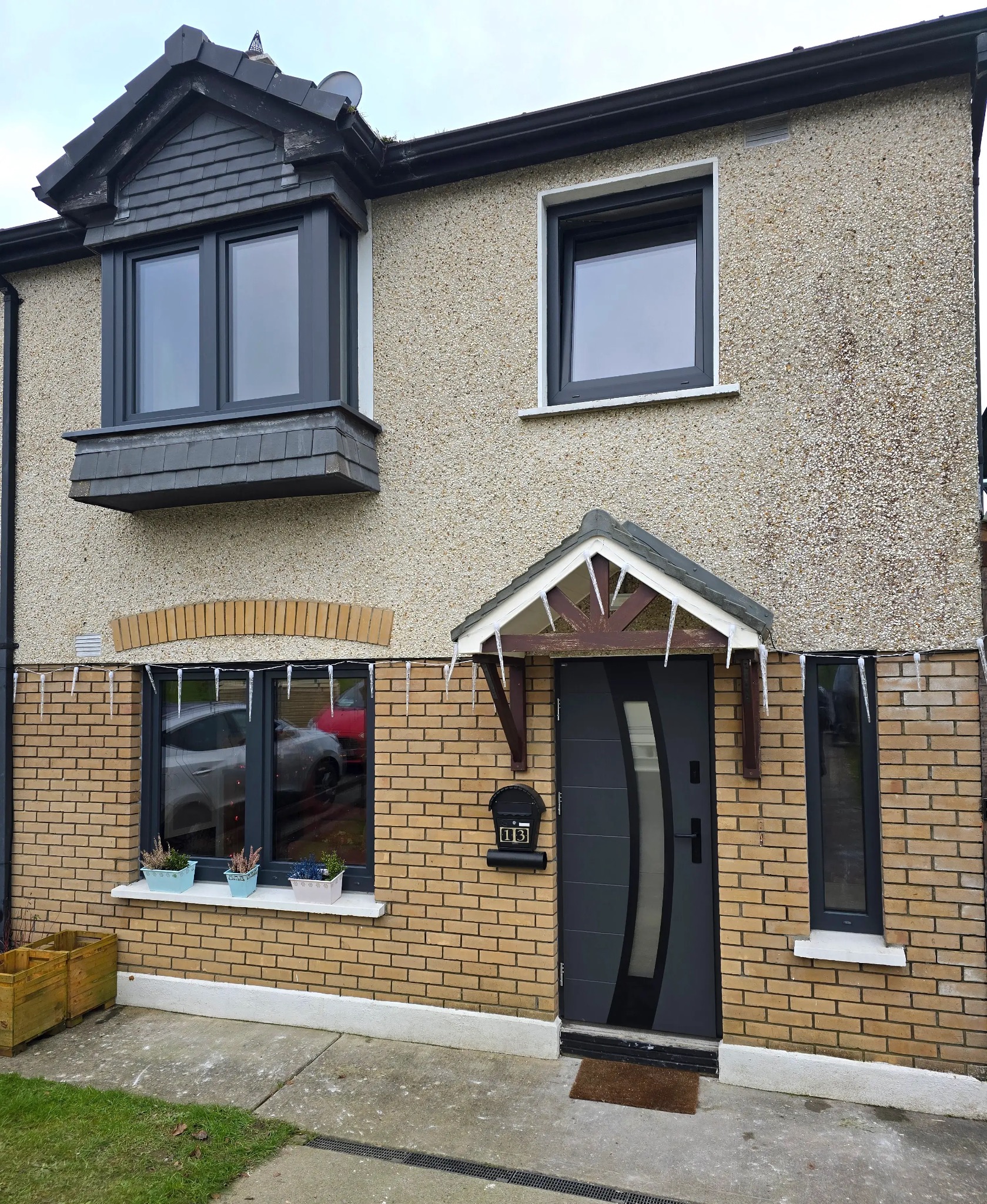 Dwukondygnacyjny dom z beżową elewacją i ceglanym parterem. Widoczne szare okna, drzwi wejściowe z daszkiem, skrzynka na listy z numerem 13 oraz kwiaty w doniczkach na parapecie okna.