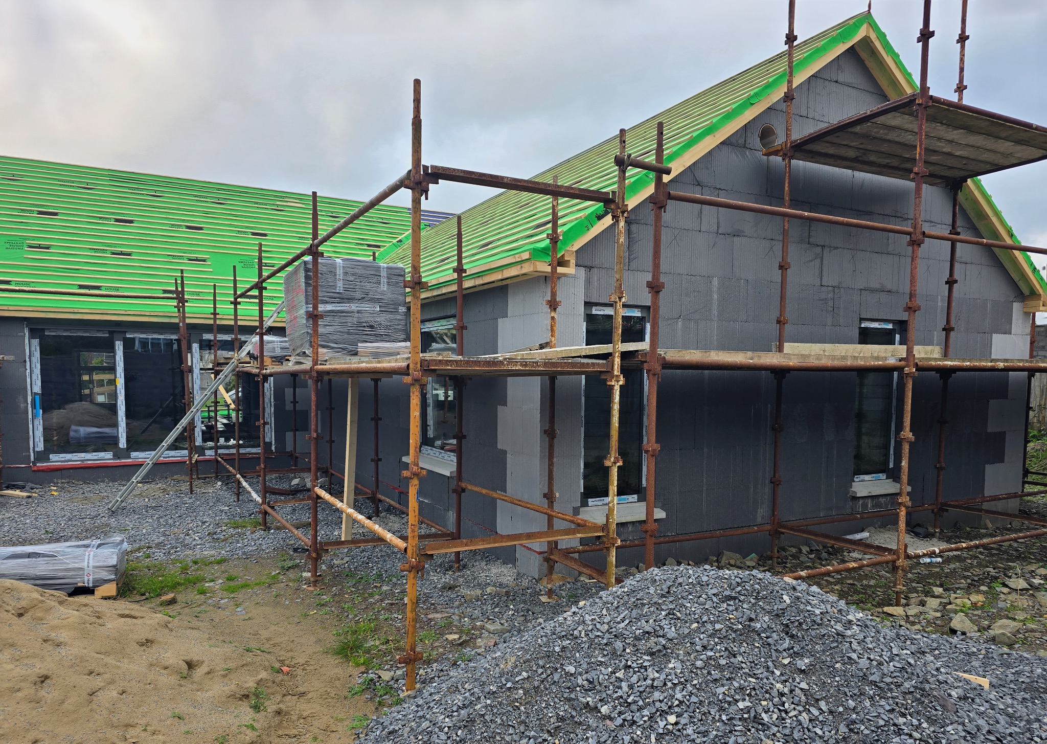 Budowa domu w trakcie ocieplania ścian zewnętrznych styropianem i montażu okien, widoczne rusztowania, materiały budowlane i zielony dach.