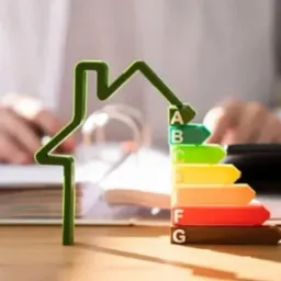 Zielony kontur domu obok kolorowej skali efektywności energetycznej A-G na biurku, osoba w tle.