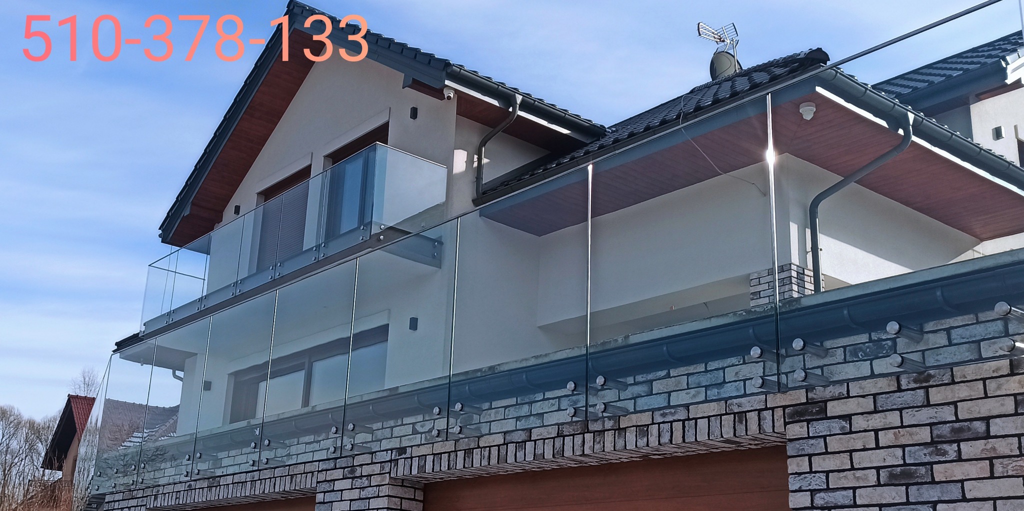 Nowoczesna dwupoziomowa balustrada ze szkła hartowanego, mocowana punktowo do ceglanej elewacji domu jednorodzinnego, widok z dołu na błękitne niebo.