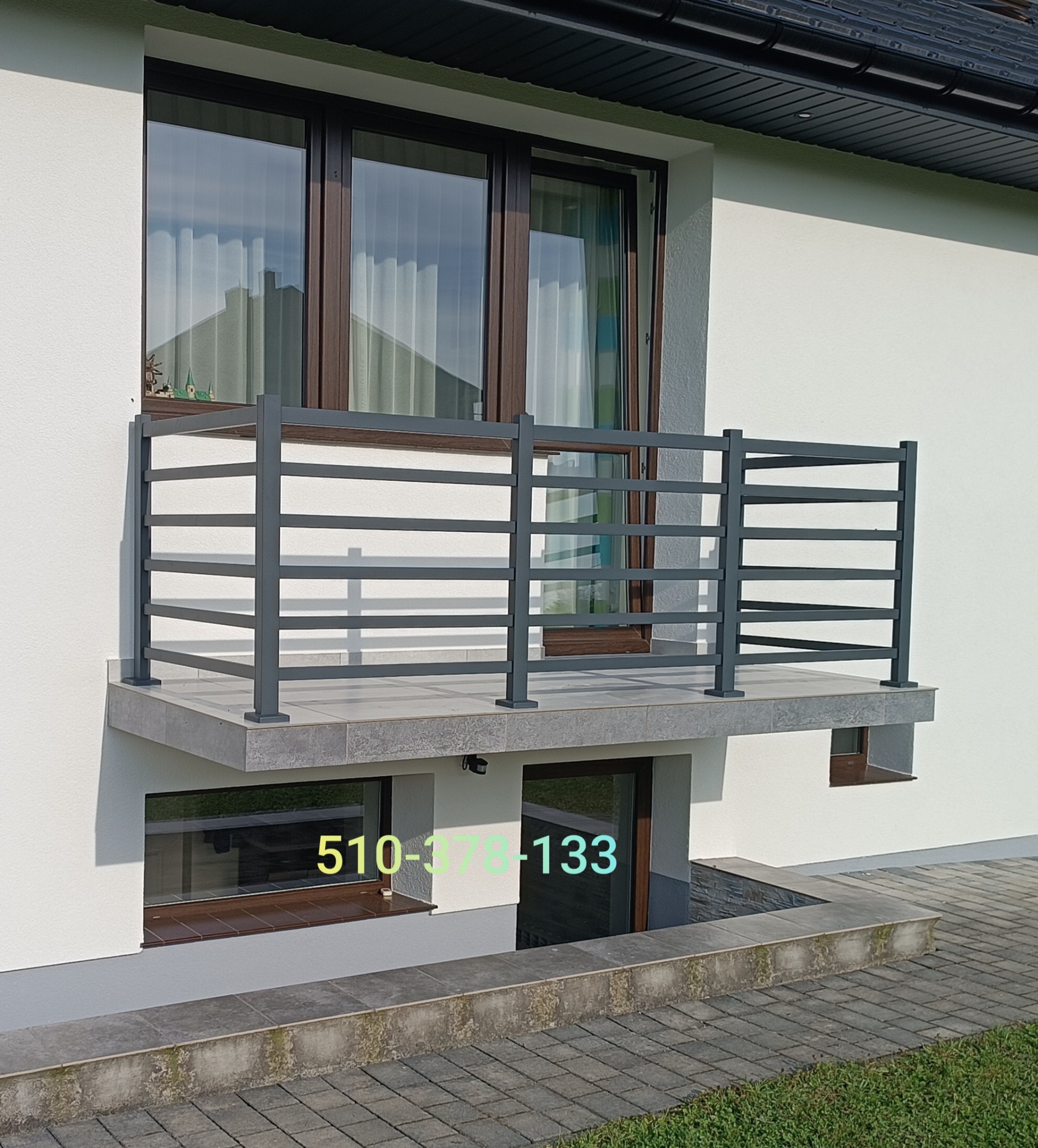 Szara, pozioma balustrada balkonowa z metalowych profili, zamontowana na balkonie z szarą płytką. W tle okna z ciemnymi ramami i fragment dachu.