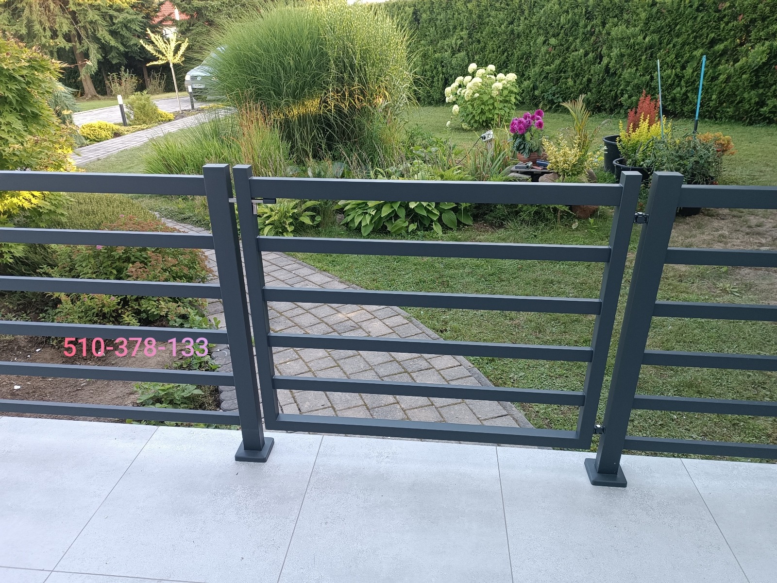 Grafitowa balustrada metalowa z poziomymi szczeblami na tle zadbanego ogrodu z kostką brukową i roślinami. Widoczny numer telefonu na balustradzie.