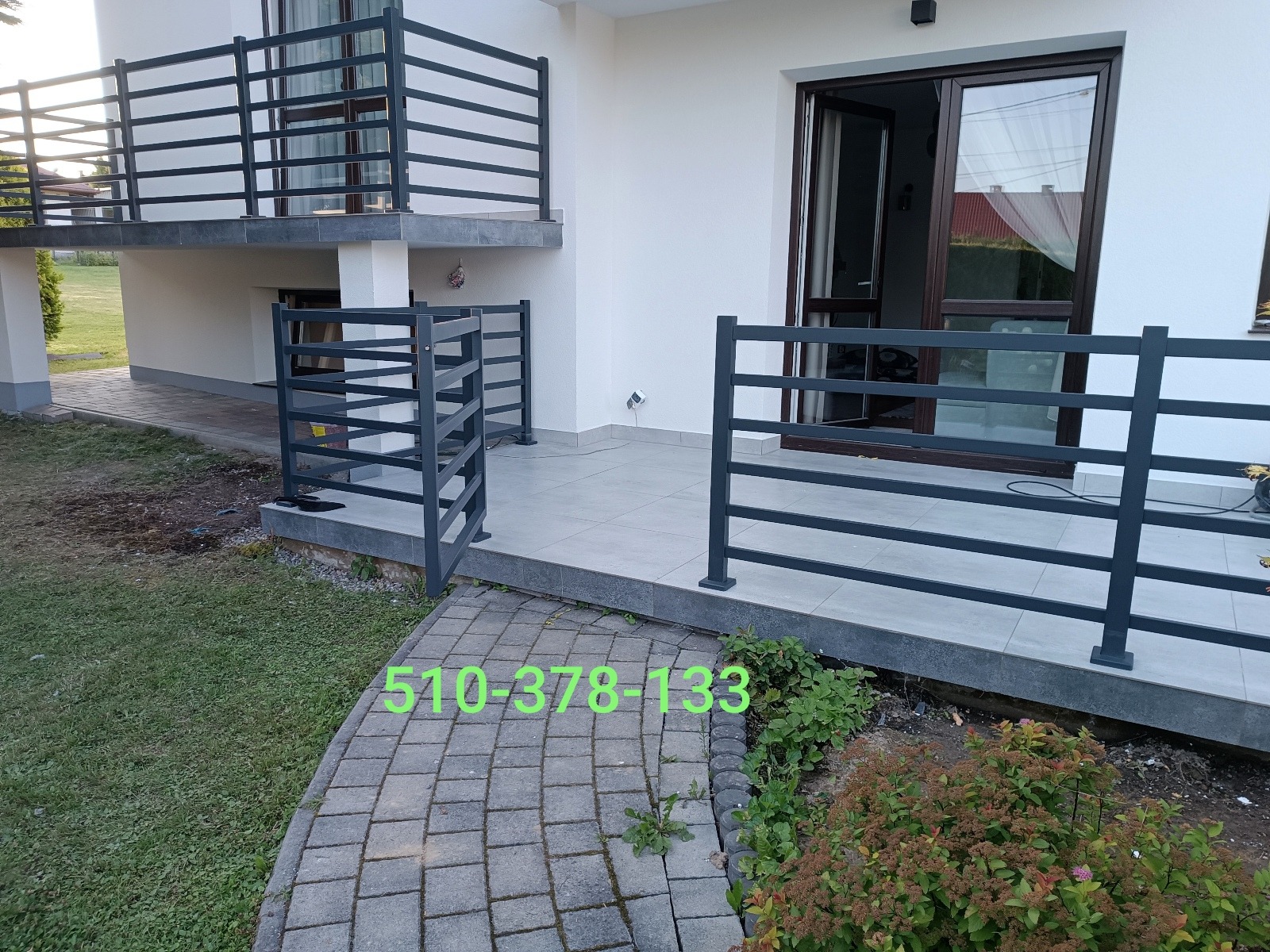 Nowoczesne, metalowe balustrady w kolorze grafitowym, zamontowane na balkonie i tarasie domu z białymi ścianami. Widoczny numer telefonu na pierwszym planie.