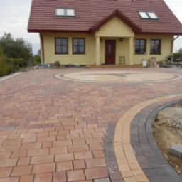 Nowo ułożona, wielobarwna kostka brukowa przed domem z okrągłym wzorem na podjeździe, widoczne krawężniki i przygotowane miejsce pod trawnik.