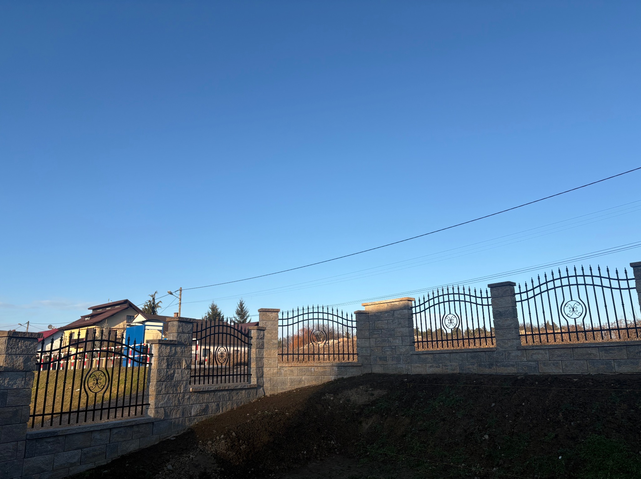 Nowoczesne ogrodzenie z czarnych, metalowych przęseł z ozdobnymi rozetami, osadzone na słupkach i podmurówce z szarej kostki łupanej, na tle błękitnego nieba i zabudowań mieszkalnych.