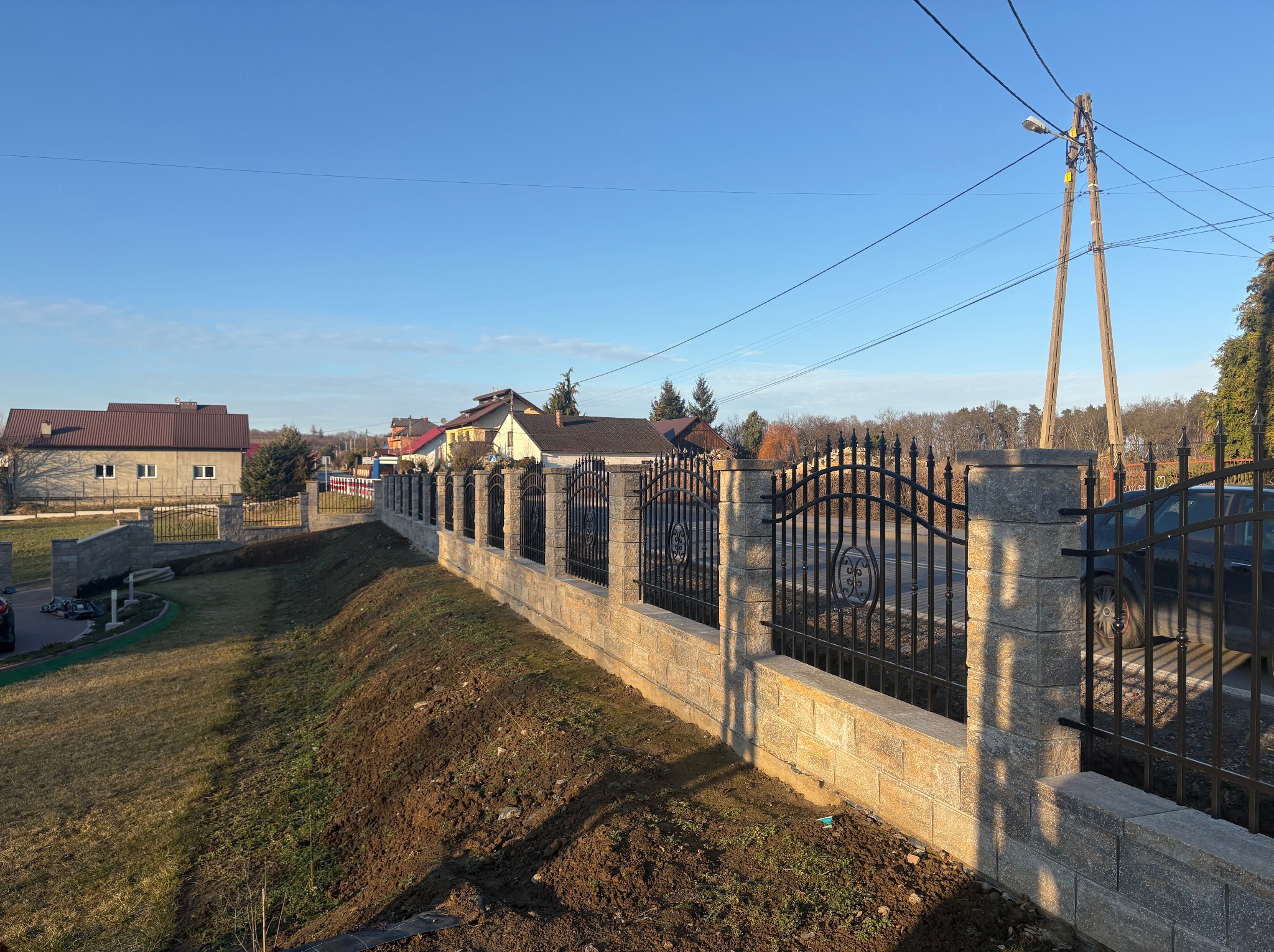 Nowoczesne ogrodzenie z kamiennymi słupkami i metalowymi przęsłami z ozdobnymi elementami, oddzielające posesję od drogi, z widocznymi domami w tle.