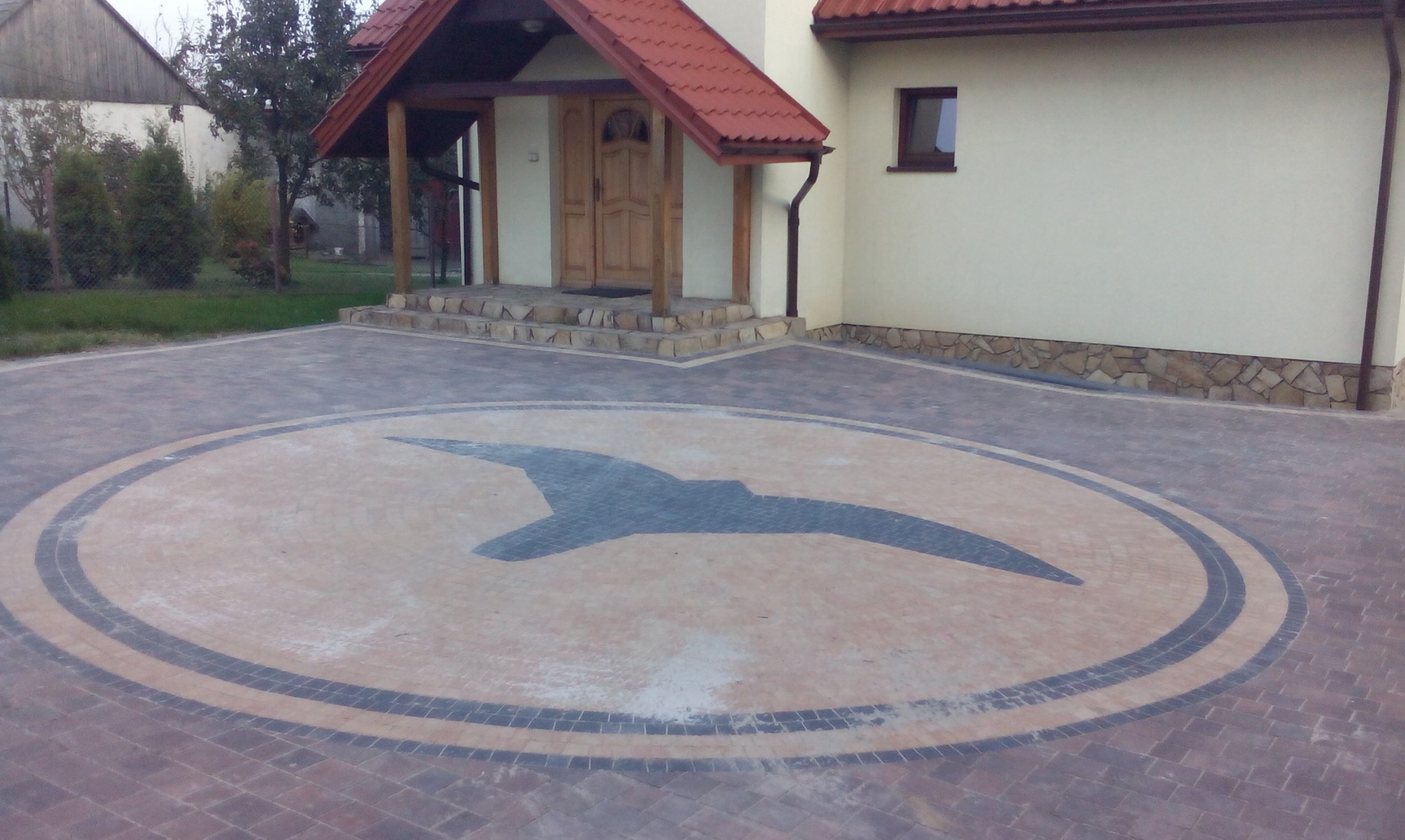 Podjazd z kostki brukowej z dekoracyjnym wzorem ptaka w centralnej części, otoczony obwódką z ciemniejszych kostek. Dom z kremową elewacją i drewnianym gankiem w tle.