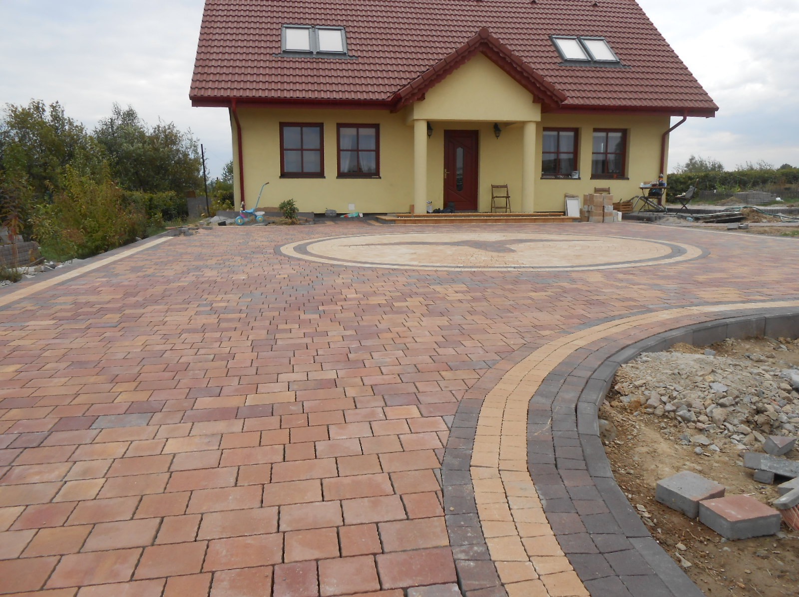 Nowo ułożona, wielobarwna kostka brukowa przed domem z okrągłym wzorem na podjeździe, widoczne krawężniki i przygotowane miejsce pod trawnik.
