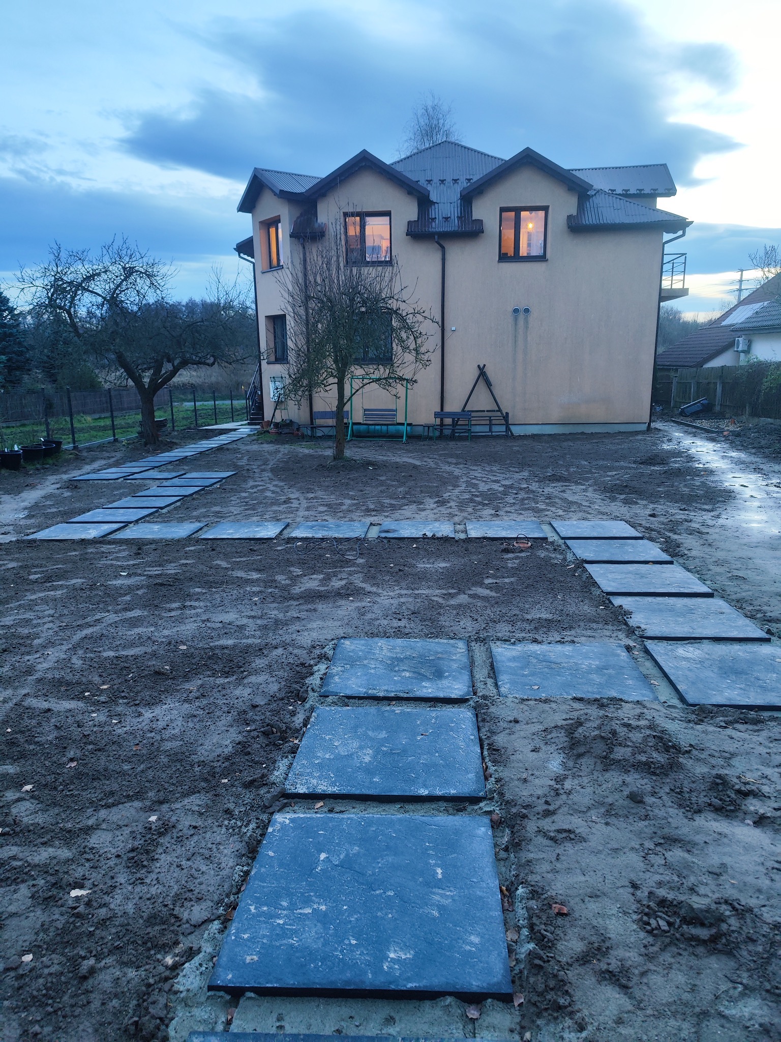 Kraków: Ułożone kwadratowe płyty chodnikowe prowadzą przez błotnisty teren w kierunku domu z podświetlonymi oknami, widok o zmierzchu.