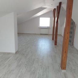 Waldemar Braczyk - Poddasze po remoncie z drewnianymi belkami, jasną podłogą imitującą drewno i białymi ścianami, okno na szczycie