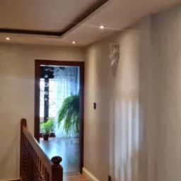 Korytarz z drewnianą podłogą i schodami, ściany w jasnym odcieniu szarości, dekoracyjny sufit z oświetleniem punktowym, widok na pokój z roślinami przez drewniane obramowanie drzwi.