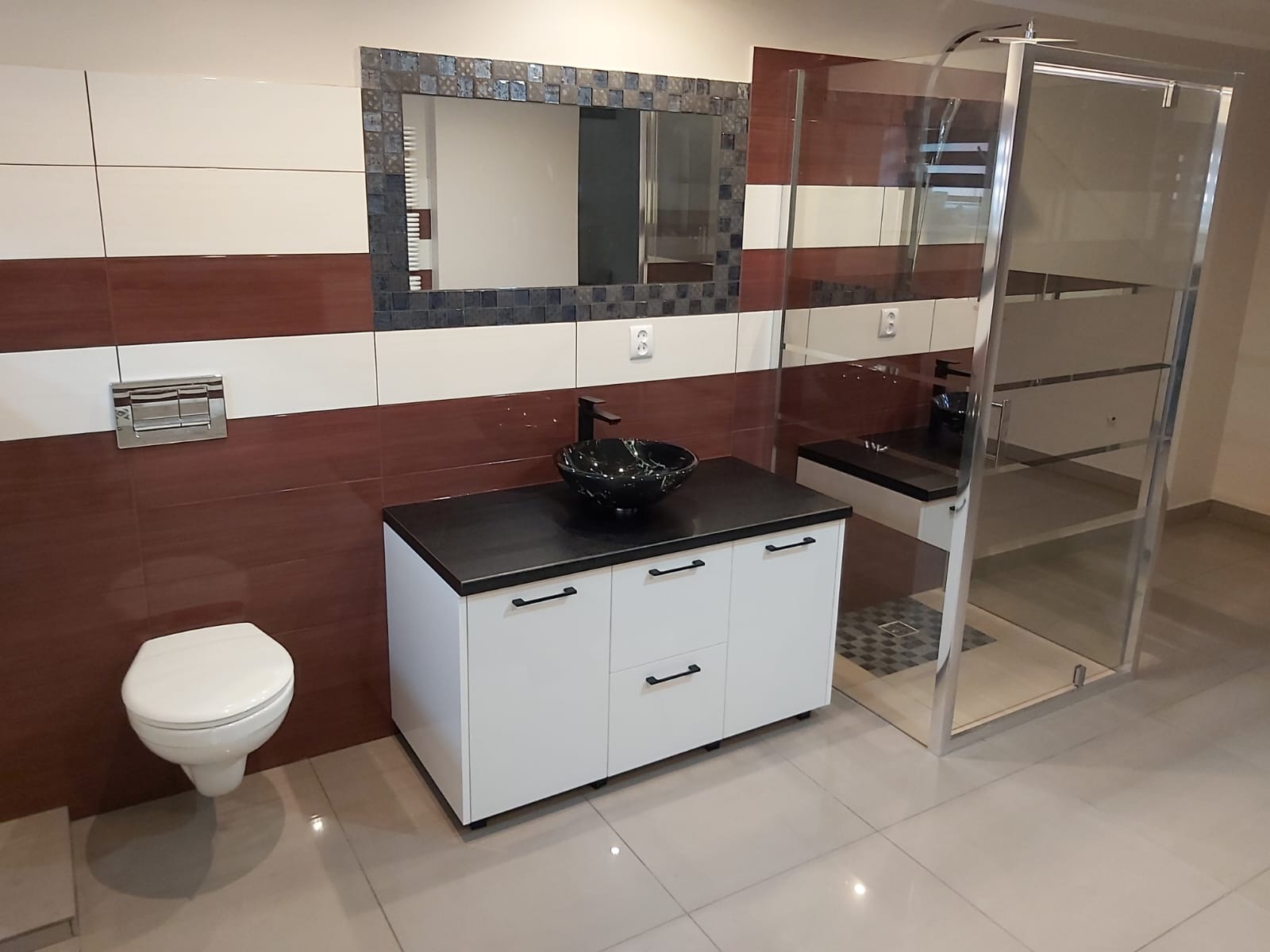 Wykończona łazienka z umywalką w kształcie misy na szafce z czarnym blatem, wiszącą toaletą i kabiną prysznicową ze srebrnymi ramami na tle płytek w odcieniach brązu i bieli oraz lustra z mozaikową...