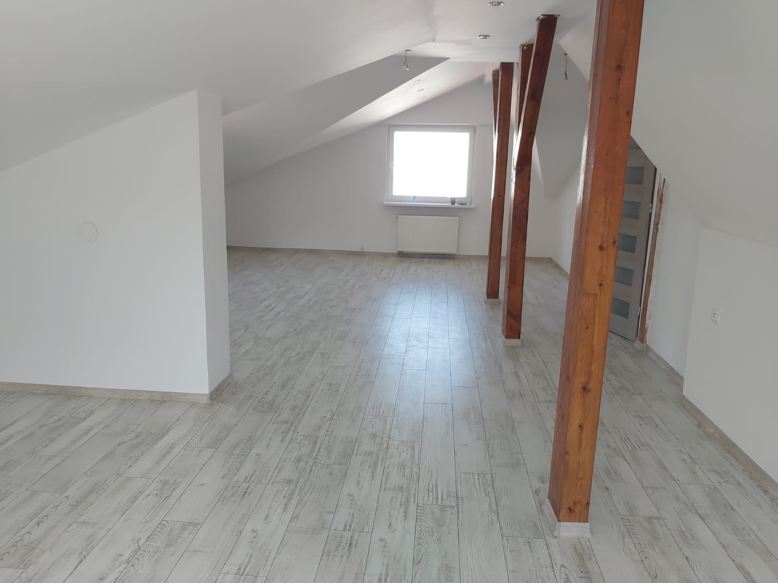 Poddasze po remoncie z drewnianymi belkami, jasną podłogą imitującą drewno i białymi ścianami, okno na szczycie