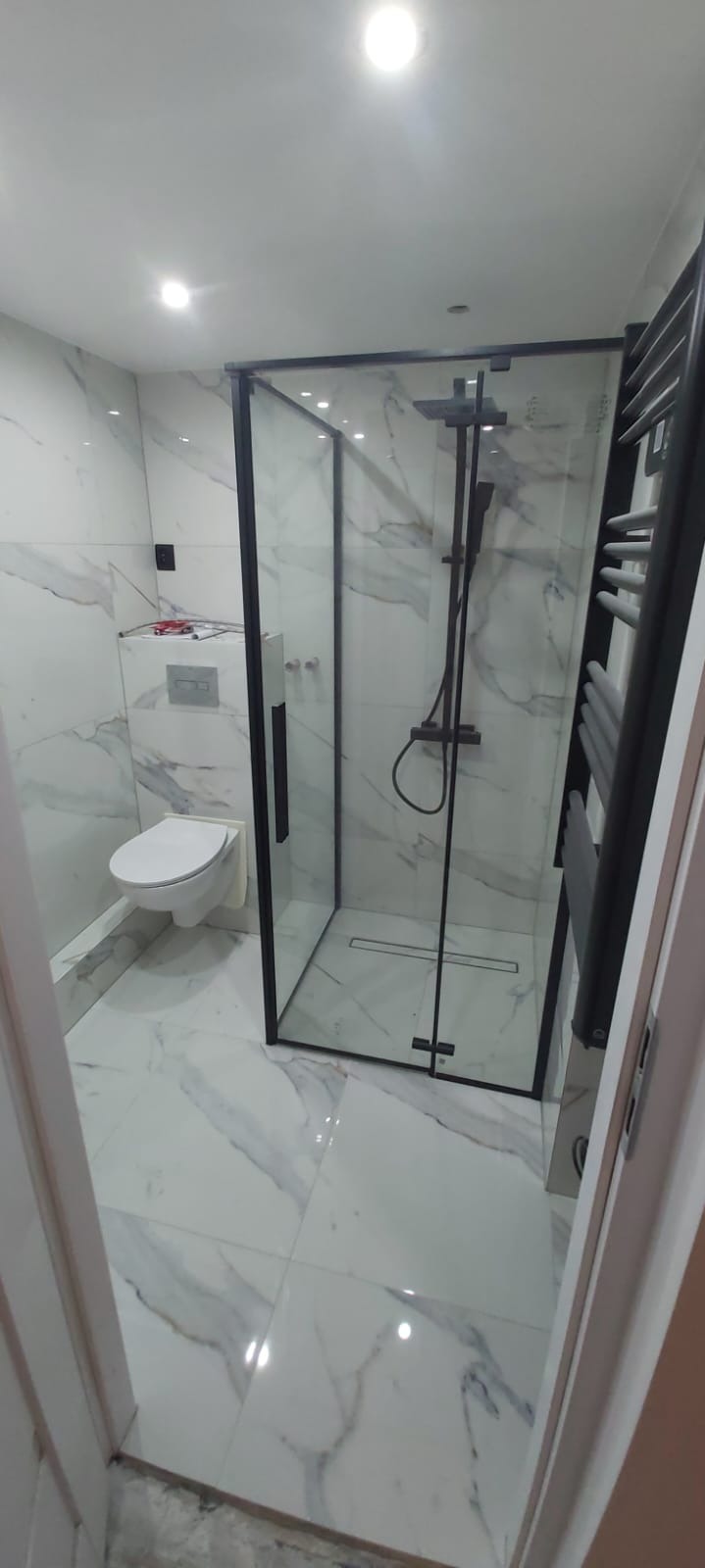 Wykończona łazienka z marmurowymi płytkami, podwieszaną toaletą i kabiną prysznicową typu walk-in z czarnymi ramami i czarnym zestawem prysznicowym. Widoczny grzejnik drabinkowy w kolorze czarnym.