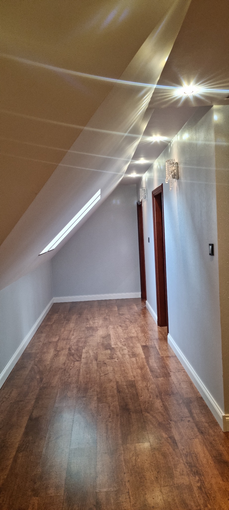 Poddasze z drewnianą podłogą, oknem dachowym i dwiema parami kryształowych kinkietów nad drzwiami.