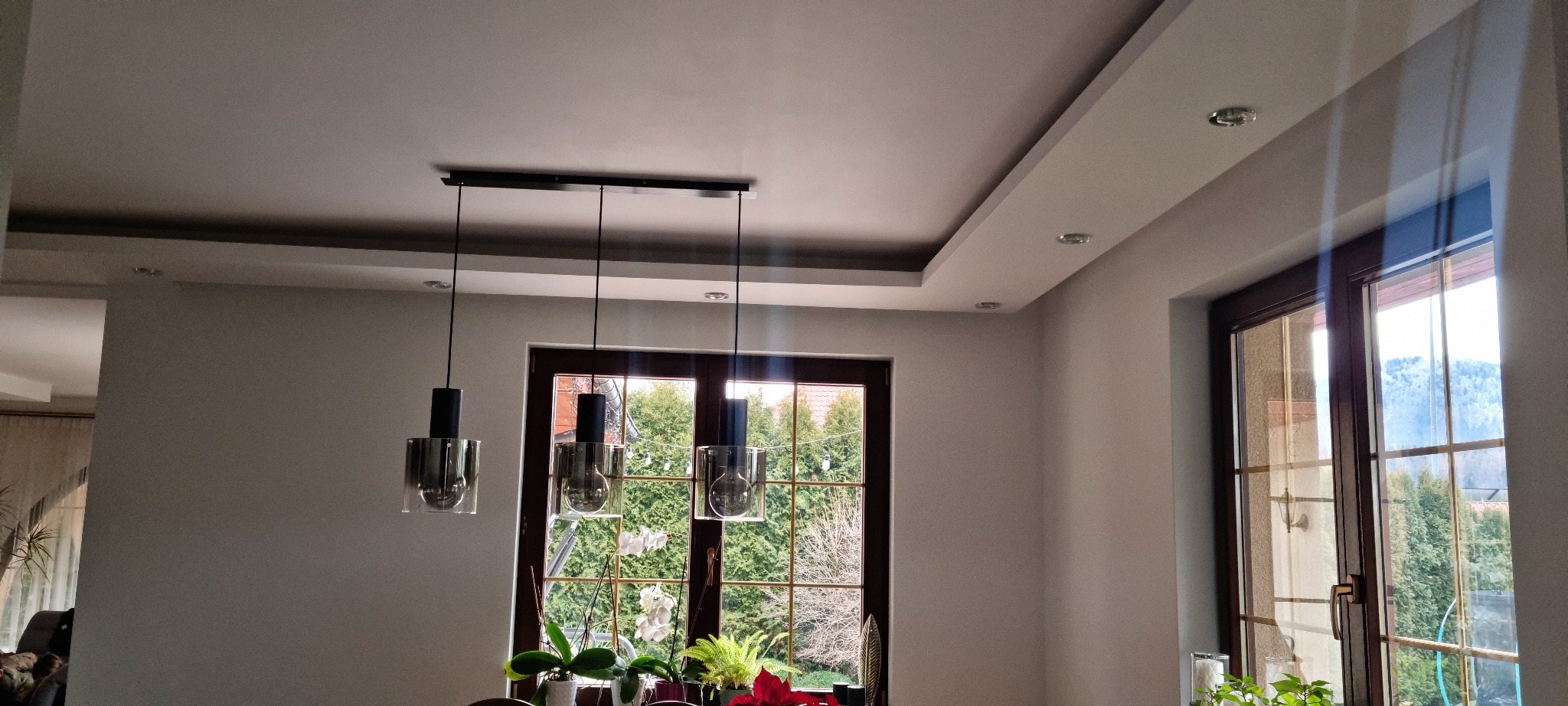 Nowoczesne oświetlenie sufitowe z trzema szklanymi lampami wiszącymi nad stołem, widok na okno z zielenią na zewnątrz, listwy dekoracyjne na suficie i oświetlenie punktowe.