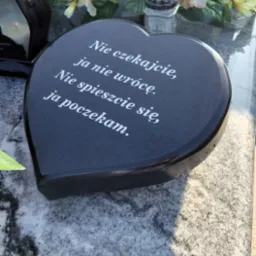 Czarny, sercowaty element nagrobny z wygrawerowanym białym tekstem, otoczony kwiatami i lampionami na szarym, marmurkowym tle