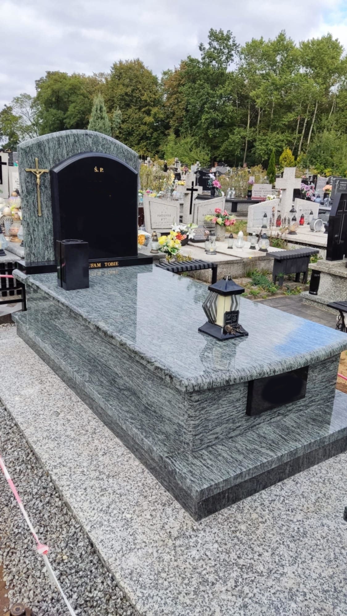 Szary granitowy nagrobek z czarną tablicą i złotym krzyżem na tle cmentarza i drzew.