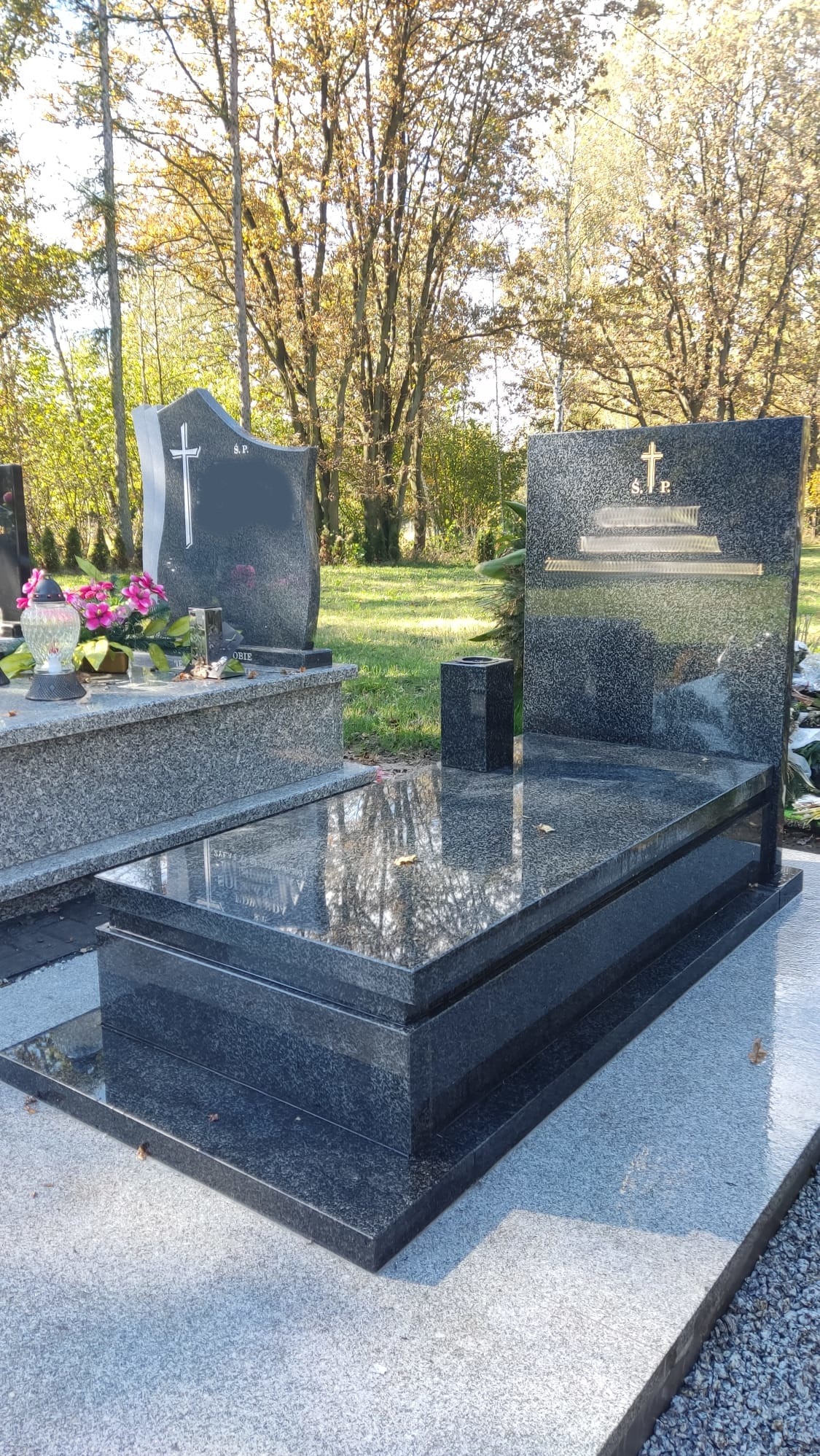 Podwójny, ciemny granitowy pomnik z inskrypcją i krzyżem na tle drzew w jesiennej scenerii.