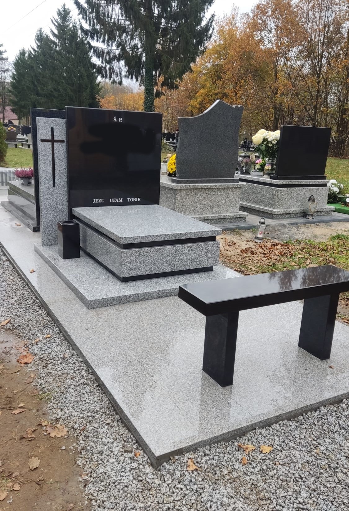 Nowoczesny nagrobek z jasnoszarego granitu z czarnymi tablicami i ławką, napis 'Jezu Ufam Tobie' na płycie nagrobnej, jesienne otoczenie cmentarza.