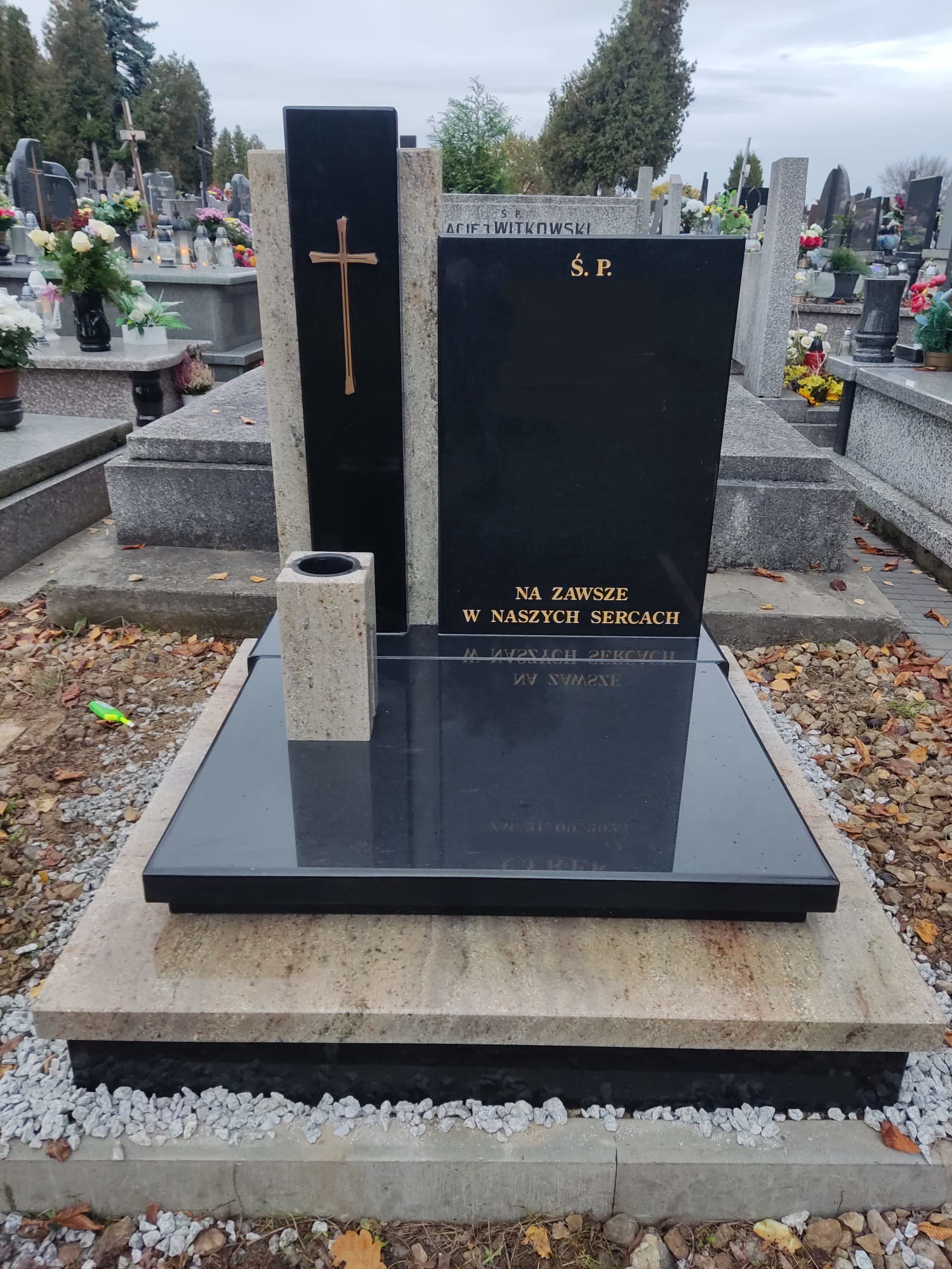 Czarny nagrobek z granitową podstawą i złotym krzyżem na cmentarzu, widoczne jesienne liście i inne nagrobki w tle.