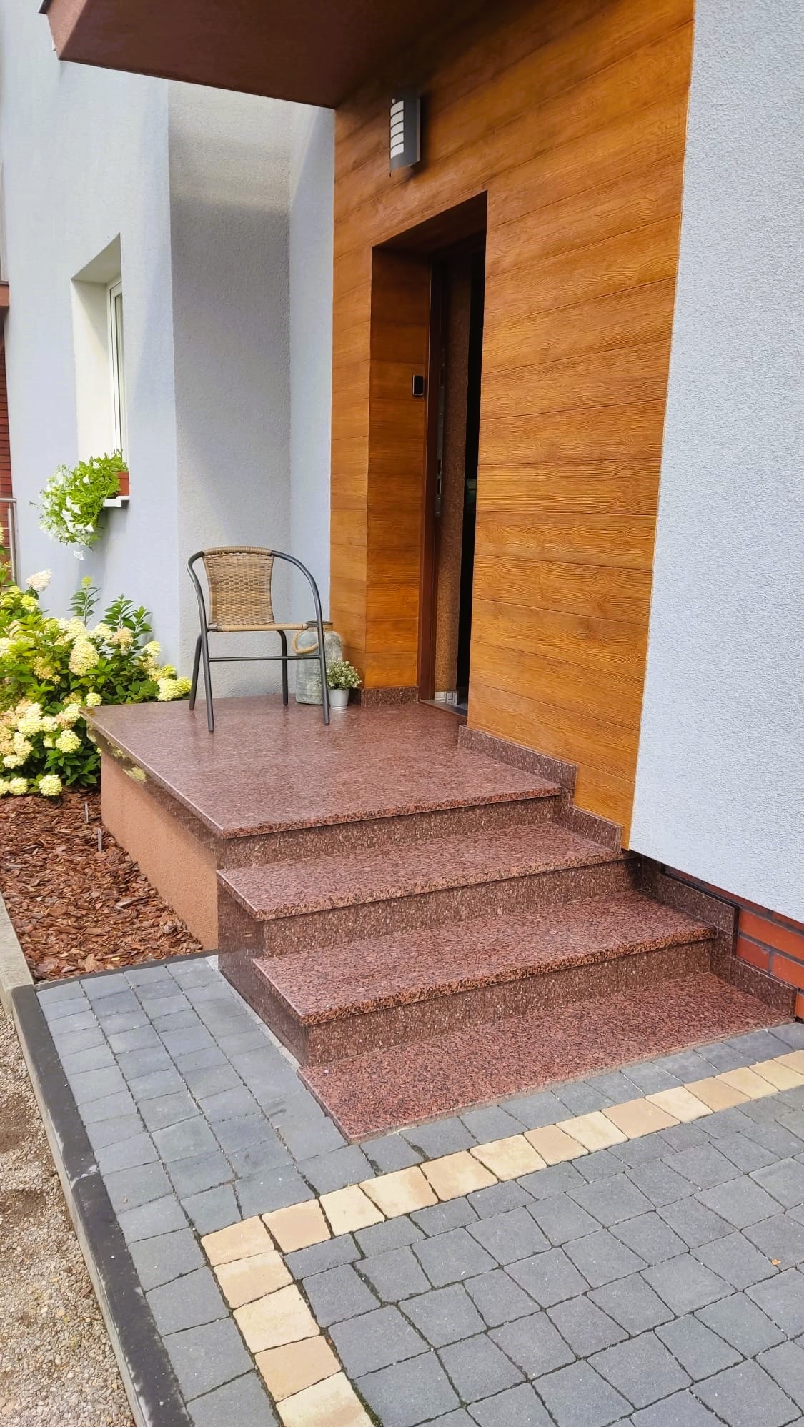 Wejście do domu z trzema stopniami z czerwonego granitu, wiklinowe krzesło z metalową ramą stoi na podeście, obok doniczka z kwiatami, drewniana okładzina ściany przy drzwiach.