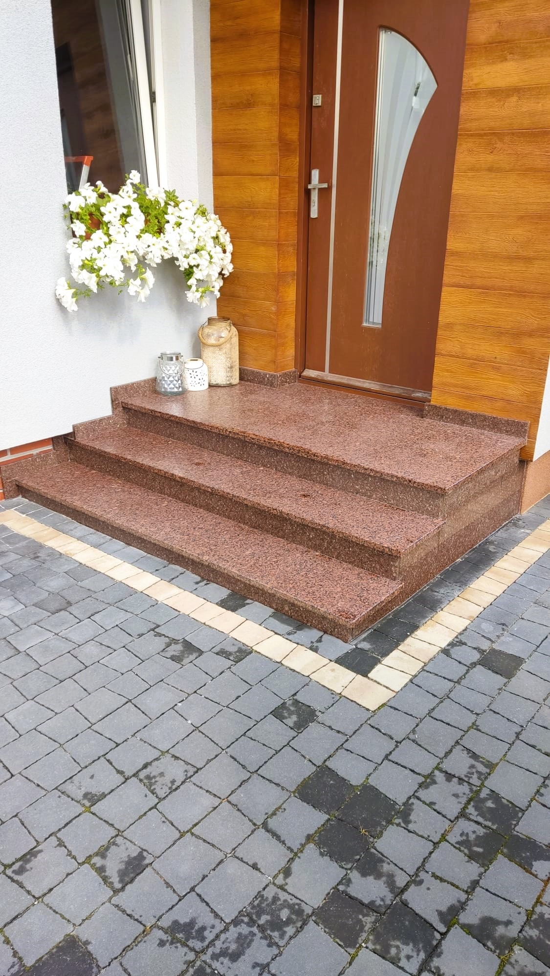 Trzy stopnie wejściowe z czerwonego granitu prowadzące do brązowych drzwi z drewnianą okładziną, ozdobione białymi kwiatami w skrzynce okiennej i dekoracyjnymi lampionami.