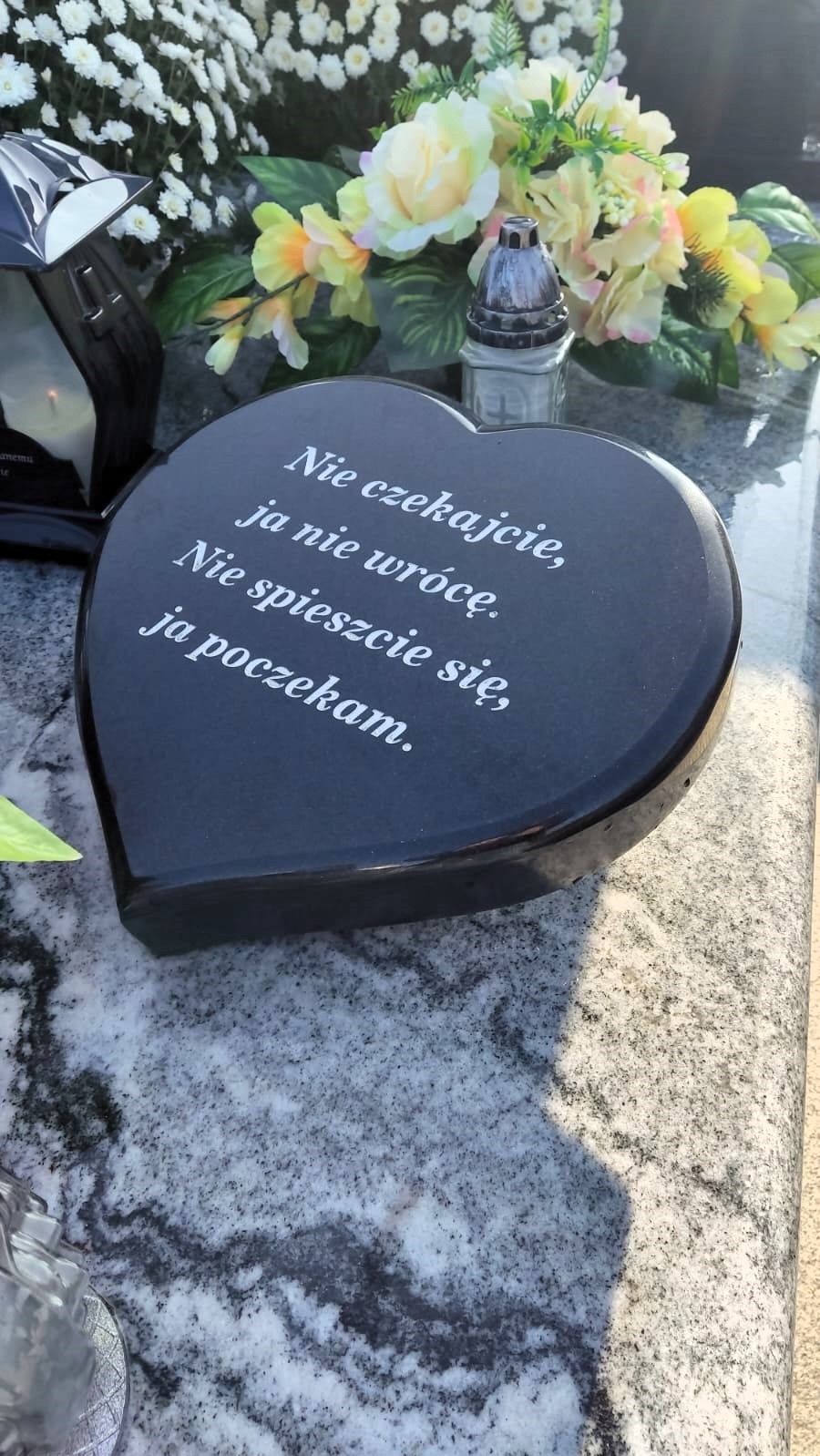 Czarny, sercowaty element nagrobny z wygrawerowanym białym tekstem, otoczony kwiatami i lampionami na szarym, marmurkowym tle