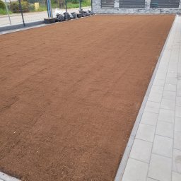 Rent-Bud-Garden KRZYSZTOF PIOTROWSKI - Starannie wyrównany, ciemnobrązowy grunt pod przyszły trawnik lub nasadzenia, z obwódką z jasnych płyt chodnikowych i workami z materiałem wzdłuż ogrodzenia.