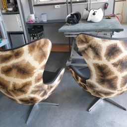 Tapicerstwo Izydorski - Dwa fotele typu Egg obite tkaniną imitującą sierść żyrafy, stojące w warsztacie, z widocznymi narzędziami i akcesoriami na stole w tle.