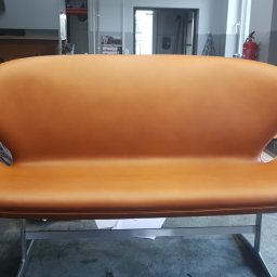 Tapicerstwo Izydorski - Dwuosobowa sofa w odcieniach karmelowej skóry na metalowym stelażu, widok z przodu w warsztacie, na podłodze leżą ścinki materiałów.