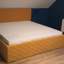 Tapicerstwo Izydorski - Żółte łóżko tapicerowane z pikowanym wezgłowiem i ramą, na nim biały materac, w pokoju z granatową ścianą i białym grzejnikiem.