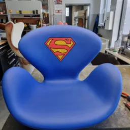 Niebieski fotel dziecięcy w kształcie litery S z logo Supermana, stojący na stole w warsztacie tapicerskim, widoczne narzędzia i maszyna do szycia w tle.