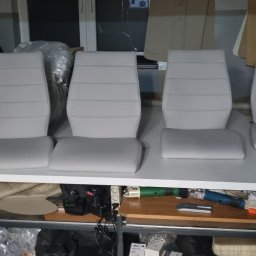 Tapicerstwo Izydorski - Cztery białe, tapicerowane siedzenia samochodowe z oparciami, ułożone obok siebie na białym stole warsztatowym, widoczne narzędzia i materiały tapicerskie w tle.