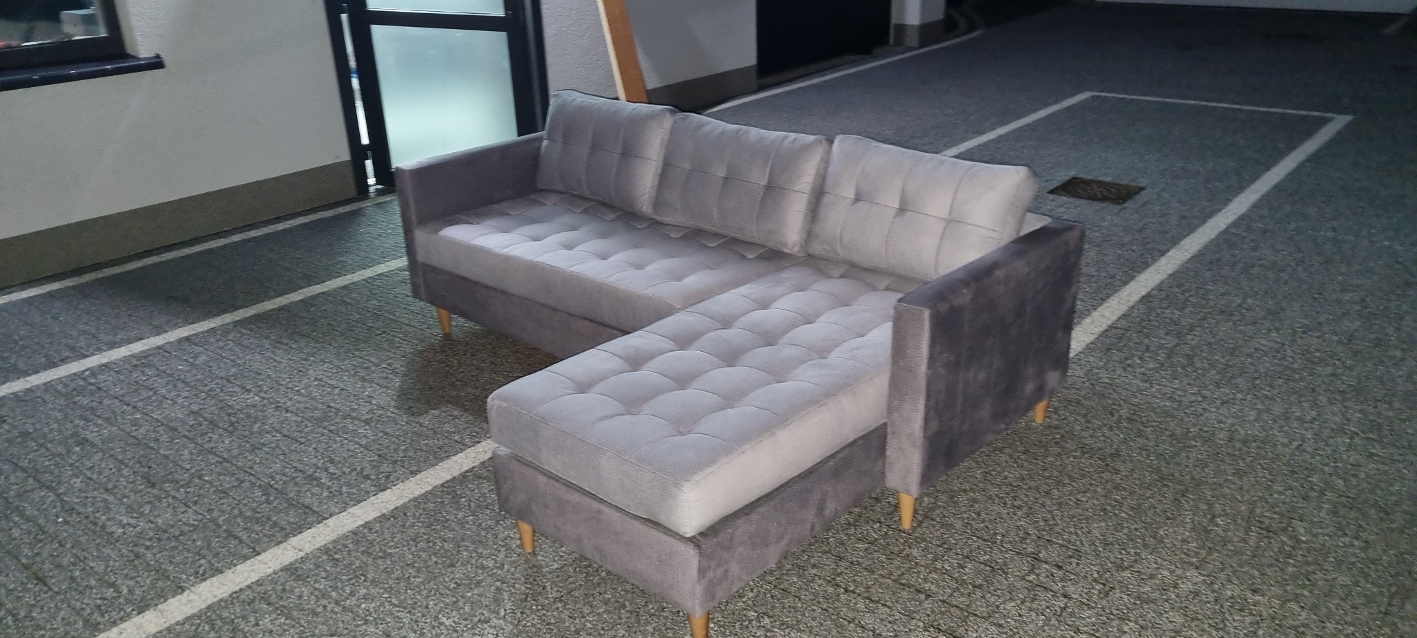 Szary narożnik z pikowanymi siedziskami i oparciami, ustawiony na zewnątrz na tle szarej nawierzchni z białymi liniami.