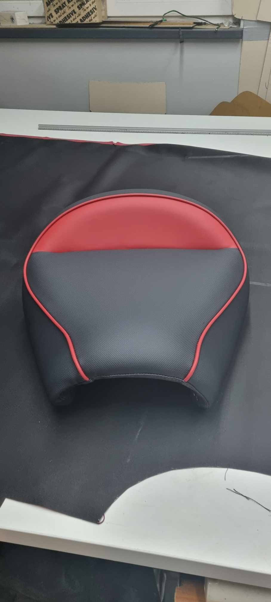 Gotowe, dwukolorowe (czerwono-szare) siedzisko z obiciem z ekoskóry, wykończone czerwoną lamówką, leżące na czarnej tkaninie na stole warsztatowym.