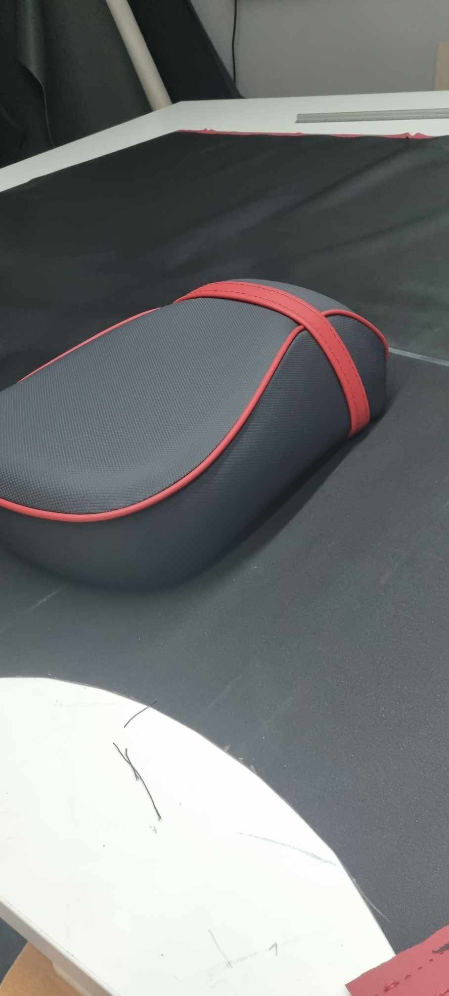 Szare siedzisko motocyklowe z czerwonymi obszyciami i pasami, leżące na czarnym materiale w warsztacie.