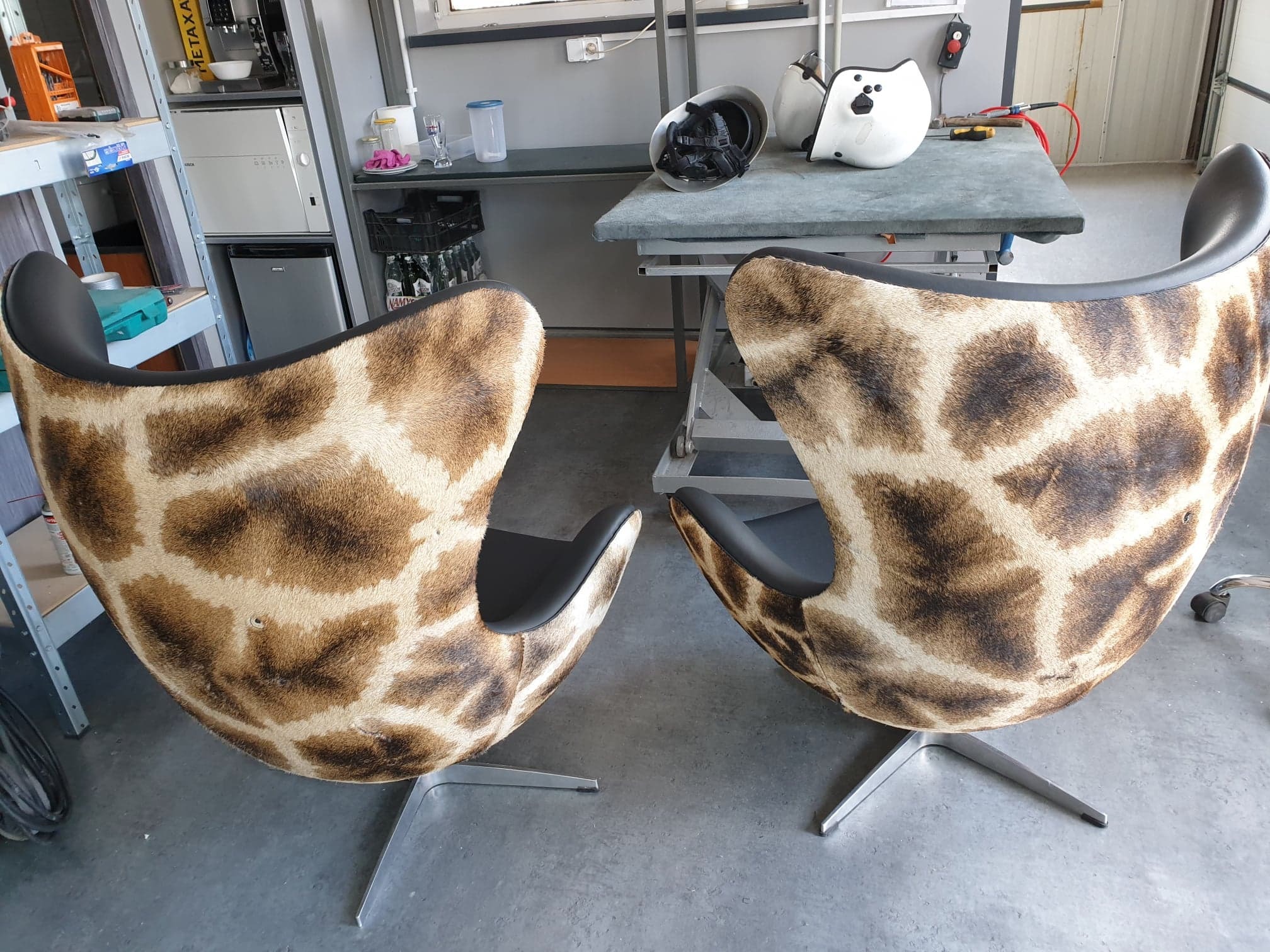 Dwa fotele typu Egg obite tkaniną imitującą sierść żyrafy, stojące w warsztacie, z widocznymi narzędziami i akcesoriami na stole w tle.