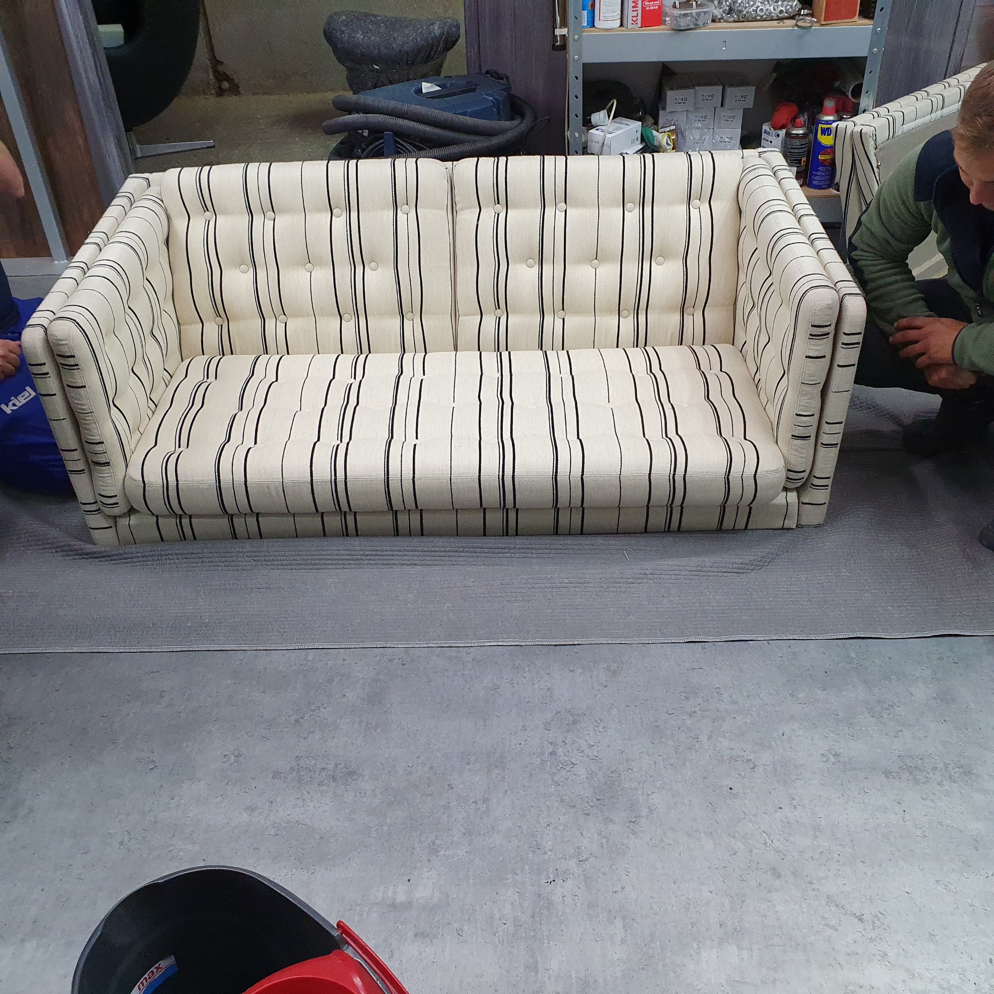 Prosty, trzyosobowy fotel z tapicerką w biało-czarne pionowe pasy, z guzikami na oparciu, ustawiony na szarej folii ochronnej w warsztacie.