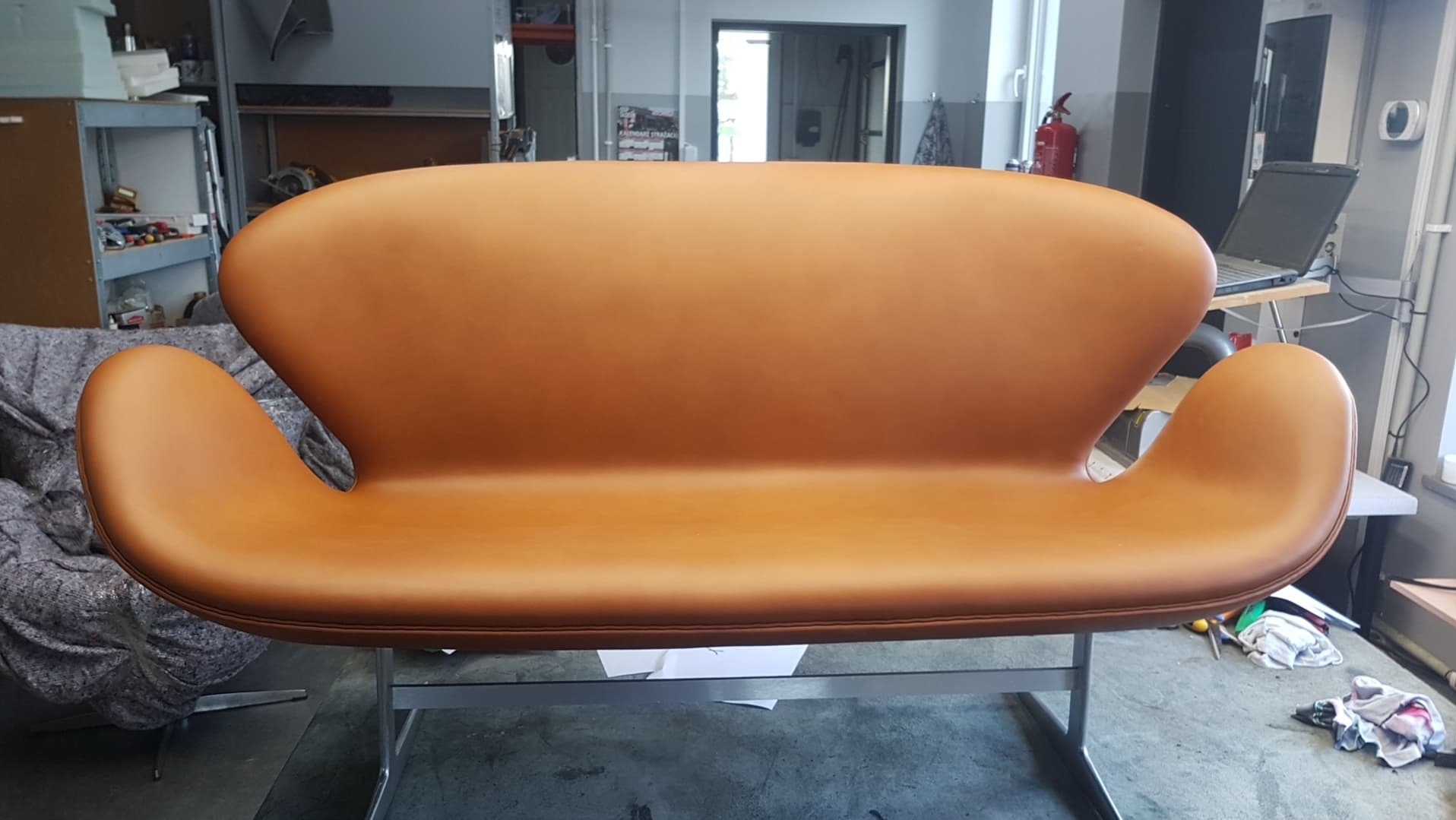 Dwuosobowa sofa w odcieniach karmelowej skóry na metalowym stelażu, widok z przodu w warsztacie, na podłodze leżą ścinki materiałów.